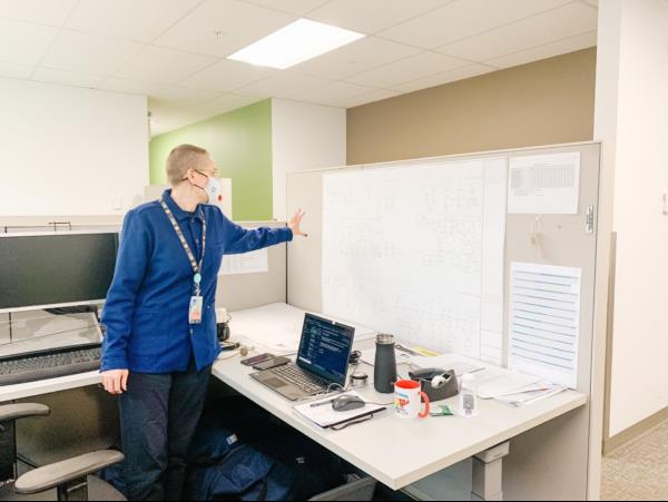 Robyn is standing at her desk looking at a technical drawing hanging on the wall of her cubicle. 