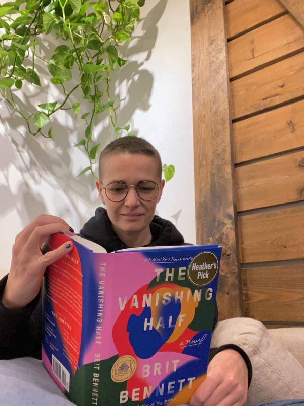 Robyn sits on the couch reading The Vanishing Half by Brit Bennett. 