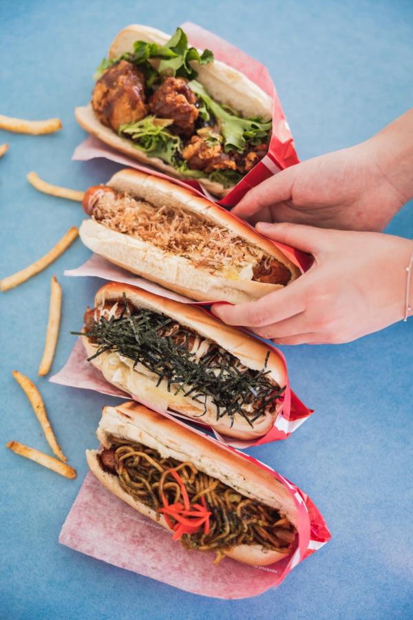 4 hot dogs on a blue backdrop, one being picked up by hand model