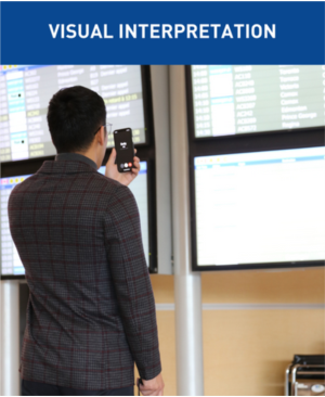 The text at the top of the image says Visual Interpretation. The photo is of a gentleman in a brown blazer holding up an iPhone in front of flight information display boards. 